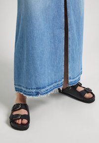 Underben iklädda en sliten blå denimkjol med snitt på sidan och svarta spännesandaler mot en vit bakgrund.