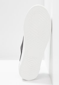 Baskets noires avec un dessus texturé et une semelle en caoutchouc blanc présentant un motif en chevron pour une meilleure adhérence. L'angle montre le détail de la partie inférieure.