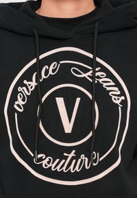 Juoda dydžio sudėtyje su dideliu šviesiai rožiniu apvaliu logotipu, kuriame yra užrašas „versace jeans couture“ ir centrinis „V“ dizainas. Su virvelėmis.