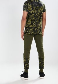 Grön t-shirt med kamouflage-mönster och korta ärmar, kombinerad med olivfärgade joggerbyxor som har kamouflage-detaljer vid mudden, tillsammans med svarta sneakers.