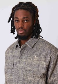 Chemise à motifs gris et vert en tissu texturé. Dotée d'un col boutonné et de deux poches poitrine. Logo sur le côté gauche.