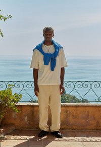 Casual antrekk med en kremfarget t-skjorte, beige bukser og en blå strikkegenser drapert over skuldrene. Sorte sko. Havutsikt i bakgrunnen.
