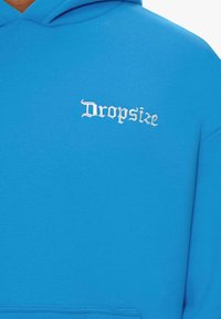 Blauer Kapuzenpullover mit weichem Material, ausgestattet mit weißen gestickten Text "Dropsize" in einer stilisierten Schriftart auf der linken Brustseite.