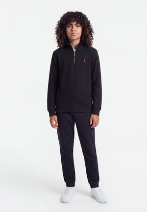 Jeune personne aux cheveux bouclés se tenant face à l’avant, portant un pull noir à fermeture éclair, un pantalon foncé et des baskets blanches sur un fond uni.