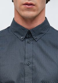 Πουκάμισο με κουμπιά σε σκούρο γκρι ύφασμα με μικρό λευκό γεωμετρικό σχέδιο, με μυτερό γιακά και δύο μαύρα κουμπιά στο μπροστινό μέρος.