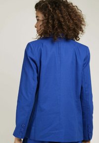 Veste bleu ajustée avec un design structuré, manches longues avec accents de boutons et texture lisse. Vue arrière mettant en avant des lignes épurées.
