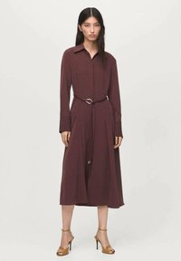 Vestito camicia lungo a maniche, di colore marrone, realizzato in tessuto liscio, con vita aderente e cintura dotata di una fibbia a forma di cuore, con un orlo di lunghezza media.