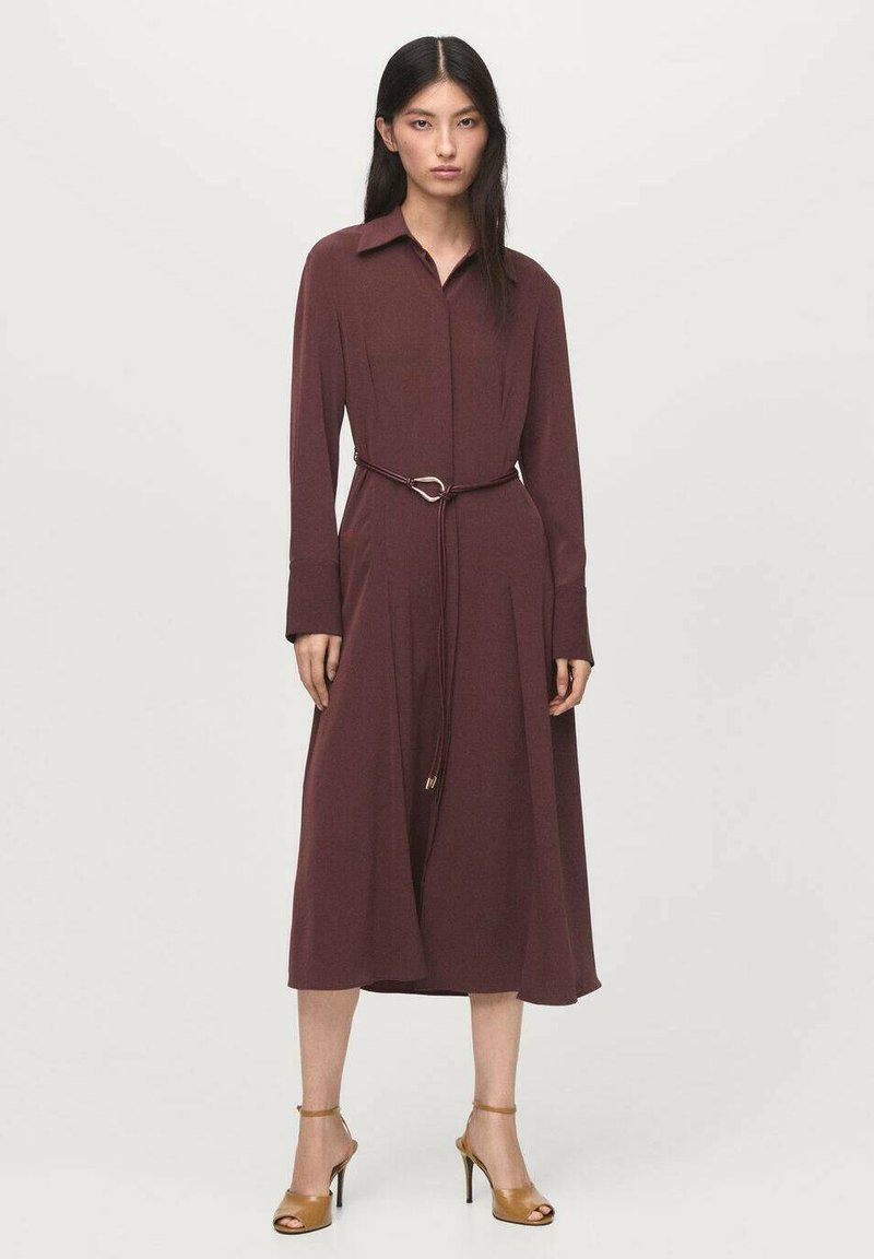 Vestito camicia lungo a maniche, di colore marrone, realizzato in tessuto liscio, con vita aderente e cintura dotata di una fibbia a forma di cuore, con un orlo di lunghezza media.