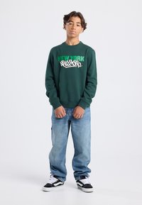 Vihreä huppari, jossa on teksti "NEW YORK" graffitityylillä, yhdistettynä vaaleansinisiin farkkuihin ja mustiin lenkkareihin, seisomassa valkoista taustaa vasten.