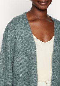 Tropisch blauwe gebreide cardigan met een open voorkant, zachte textuur en verlaagde schouders, gelaagd over een lichtbeige v-hals top.