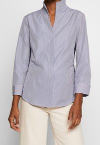 Randig blus i marinblått och vitt, med hög krage, långa ärmar och rundad nederkant. Tillverkad av lätt, slät tyg.