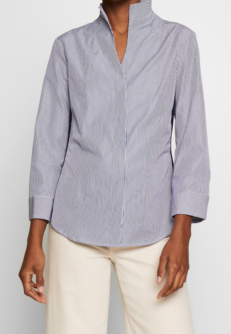 Randig blus i marinblått och vitt, med hög krage, långa ärmar och rundad nederkant. Tillverkad av lätt, slät tyg.