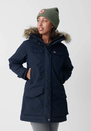 Marineblauwe parka met een bont-gevoerde capuchon, voor knopen en twee zijzakken. Het materiaal is soepel met een lichte glans. Geribbelde groene beanie.