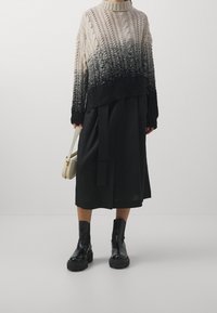 Pull en tricot avec un dégradé de crème à noir, présentant un ourlet et des poignets côtelés. Associé à une jupe midi noire et des bottes à talons noires.