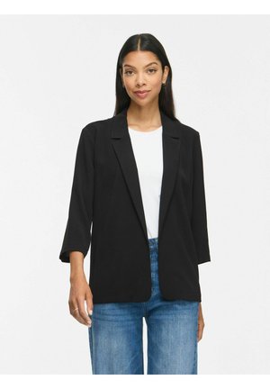 Kvinde med langt mørkt hår iført en sort blazer, hvid skjorte og blå jeans, stående mod en ensfarvet lyse baggrund.
