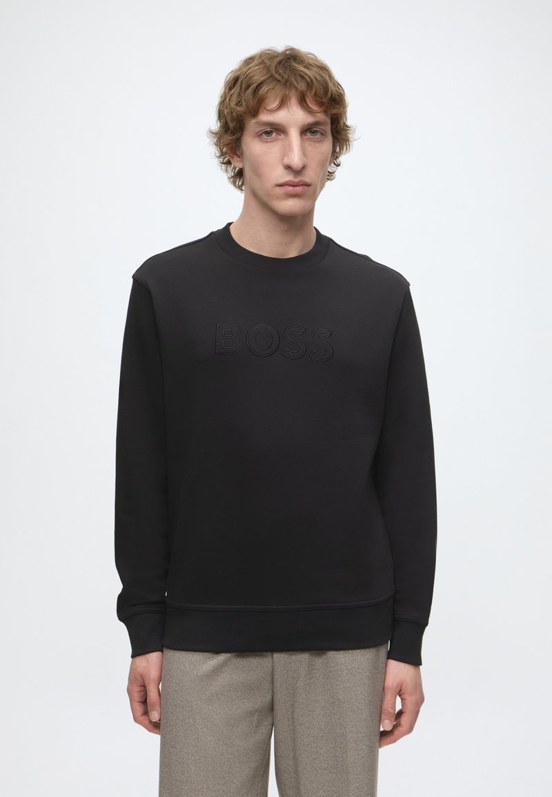 Jeune homme aux cheveux bouclés portant un sweat-shirt noir avec logo BOSS en relief et un pantalon gris, debout devant un fond clair uni.