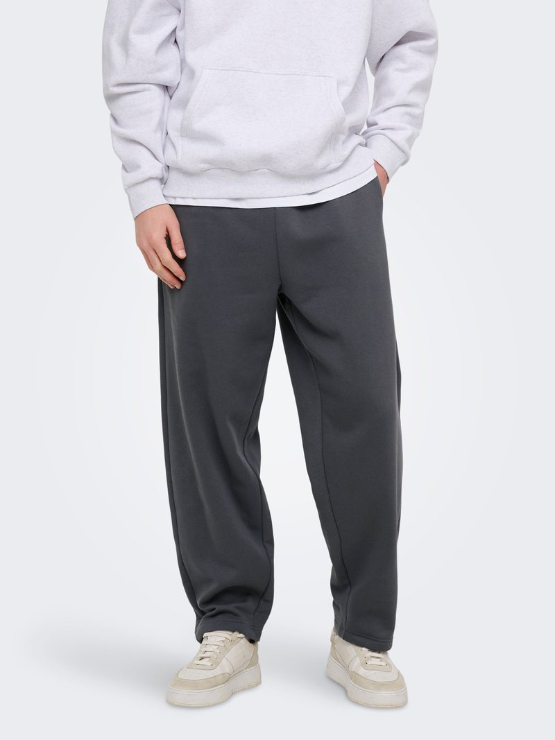 Oseba, oblečena v temno sive sproščene športne hlače, bele superge in svetlo sivo jopa s kapuco, z rokami v žepih, stoji na belem ozadju.