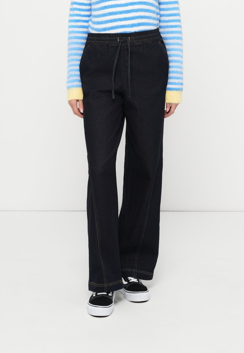 Temno modre jeansa z ohlapnim krojem, elastičnim pasom in vidnimi šivi. Kombinirane z modro-rumeno črtastim puloverjem.