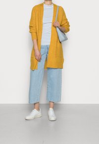 Cardigan lungo giallo senape, camicia a righe bianche e blu, jeans a gamba larga blu chiaro e sneakers bianche. Borsa grigia con accenti in argento.