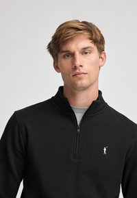 Svart quarter-zip sweatshirt med en slät yta, med en liten vit logotyp på vänster bröst och en grå tröja under.