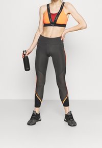 Czarne legginsy sportowe z pomarańczowymi akcentami, w zestawie z wielokolorowym stanikiem sportowym. Trzyma czarną butelkę na wodę ze stali nierdzewnej.