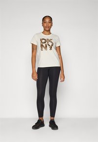 Krémové tričko s leopardím potlačou loga "DKNY", v kombinácii s čiernymi legínami a čiernymi teniskami. Hladká tkanina, pohodlný strih.