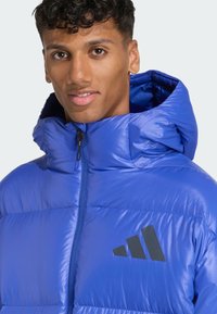 Modrá prešívaná bunda má vysoký golier, plný zips a čierne logo na hrudi. Materiál je lesklý a izolačný, aby poskytoval teplo.