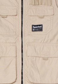 Beige Timberland-väst med dragkedja framtill, fyra stora fickor och varumärkesetikett på övre vänstra sidan. Tyget har en strukturerad väv.