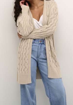 Cardigan beige a maglia con apertura frontale, caratterizzato da un motivo tessurizzato e polsini a coste. Abbinato a jeans azzurri a vita alta.