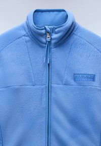 Giacca in fleece blu chiaro con colletto alto, cerniera frontale e tessuto testurizzato. Presenta una patch con logo nella parte superiore sinistra.