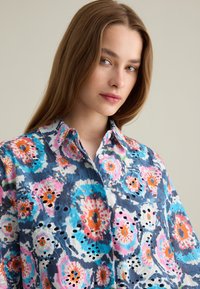 Camicia a maniche lunghe con stampa floreale caratterizzata da motivi blu, rosa e arancione, realizzata in tessuto leggero con fori a occhio e colletto a bottoni.
