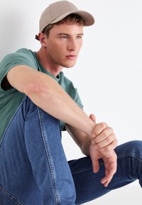 Gorra beige clara, camiseta verde de cuello redondo y jeans de mezclilla azul. Modelo sentado con los brazos descansando sobre las rodillas, mostrando las texturas y colores de las telas.