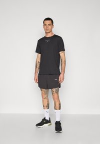 Sort t-shirt med logo, sammen med mørke shorts. Har tatoveringer på armene, hvide sokker og sorte sneakers med grønne detaljer.