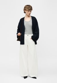 Γυναίκα με κοντά μαλλιά που φοράει γκρι αμάνικο τοπ, μαύρη φαρδιά ζακέτα, φαρδιά λευκά παντελόνια, μαύρες ψηλοτάκουνες γόβες και κρατάει σκοτεινή τσάντα ώμου.