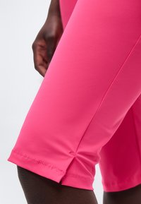 Zbliżenie na osobę o ciemnej karnacji, ubraną w jasnoróżowe dopasowane szorty, z widocznym szczegółowym przeszyciem i małym bocznym rozcięciem przy brzegu.