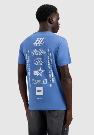 Man draagt een blauw T-shirt met verschillende witte "Ballin"-logo's en tekst op de achterkant, gecombineerd met donkergrijze jeans, zijwaarts gekeerd.