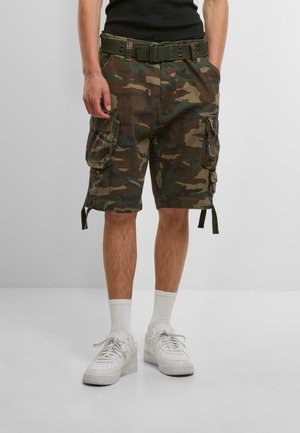 Camo Cargo-Shorts in Grün und Braun mit mehreren Taschen, einem Gürtel und seitlichen Riemen. Getragen mit weißen Sneakers und Kniestrümpfen.