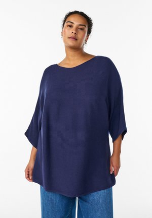 Navy blue oversized top with a ribbed texture, wide neckline, and batwing sleeves, paired with light blue denim jeans.