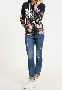Sudadera con capucha floral de base azul marino, cordones blancos y logo. Combinada con jeans azul desgastados y zapatillas blancas.