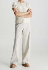 Beige bomullströja med "Calvin Klein"-logotyp, parats med ljusfärgade vida byxor och bruna plattformssandaler med flätade remmar.