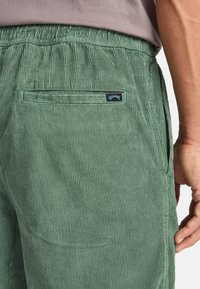 Green corduroy pants featuring a textured surface, elastic waistband, and a back patch pocket with a blue label.