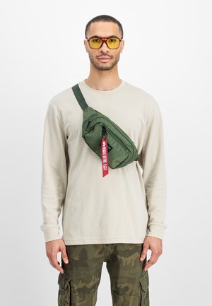 Grønn nylon crossover-veske med et elegant design, en glidelåslomme og en rød merkelapp, båret over en beige langermet skjorte og kamuflasjebukser.