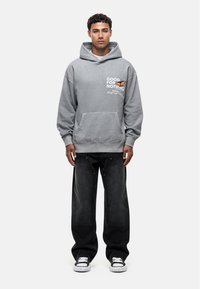 Felpa grigia con grande cappuccio, tasca a marsupio e grafica a farfalla. Indossata con pantaloni neri a gamba larga e sneakers grigie. Design casual.