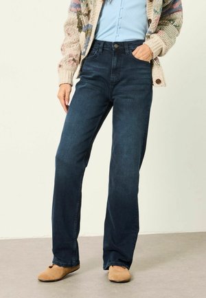 Persoon die donkerblauwe high-waist rechte spijkerbroek draagt, lichtblauwe top met knopen, gebreide cardigan met patroon en beige platte schoenen, staand met één hand in de zak.