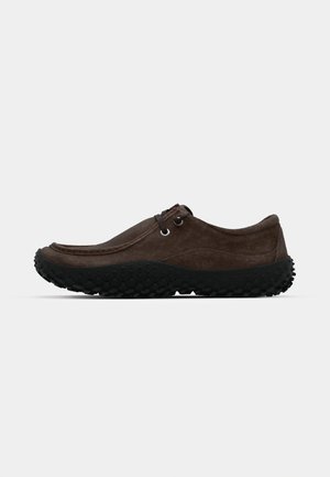 Zapatos de ante marrón con puntera redondeada, suela de goma negra con un patrón de agarre texturizado y un diseño minimalista. Sin cordones visibles.