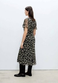 Schwarzes Blumenkleid mit durchsichtigen Ärmeln, das einen fließenden Rock und eine taillierte Taille hat. Das Modell trägt schwarze Stiefel. Rückansicht dargestellt.