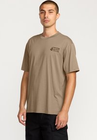 T-shirt beige clair à manches courtes avec un col rond et un petit logo noir imprimé sur le côté supérieur gauche. Matière en coton.