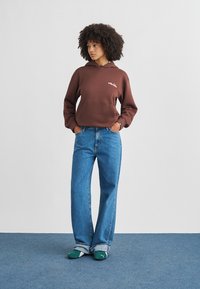 Femme aux cheveux bouclés portant un sweat à capuche marron, un jean bleu ample et des chaussons Puma verts, debout les mains dans les poches sur un tapis bleu.