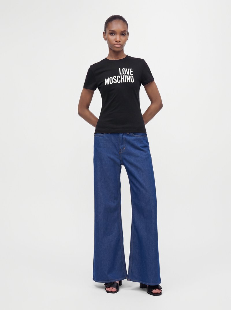 Жена, носеща черна тениска "Love Moschino", широки сини дънки и черни обувки с отворени пръсти, стои с ръце зад гърба си.