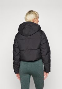 Veste courte matelassée noire avec capuche, présentant des coutures horizontales et une texture lisse et brillante, portée sur des leggings verts.